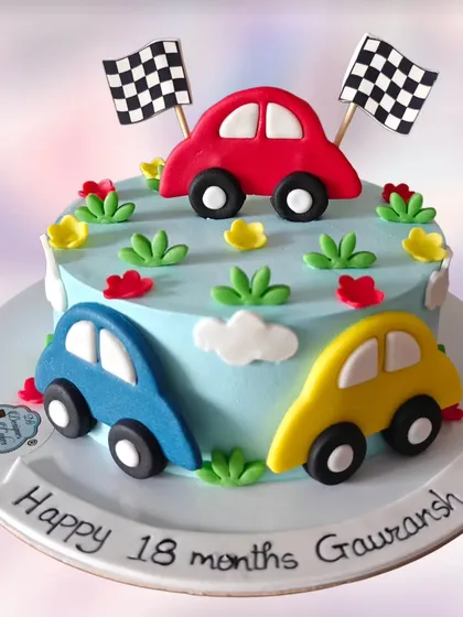 A simple and sweet car-themed cake for an 18-month celebration. It features three colorful fondant cars on a light blue whipped cream base.