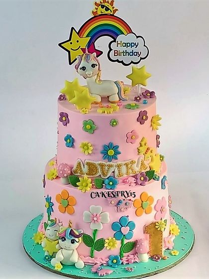 A cheerful two-tier unicorn and rainbow cake. This pink cake is covered in colorful fondant flowers and features cute unicorn figures for a happy first birthday.