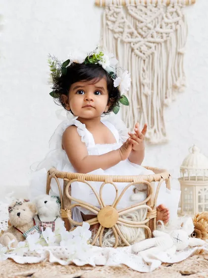 A serene portrait of a baby girl in a white dress and floral crown, posing elegantly in a wicker basket.