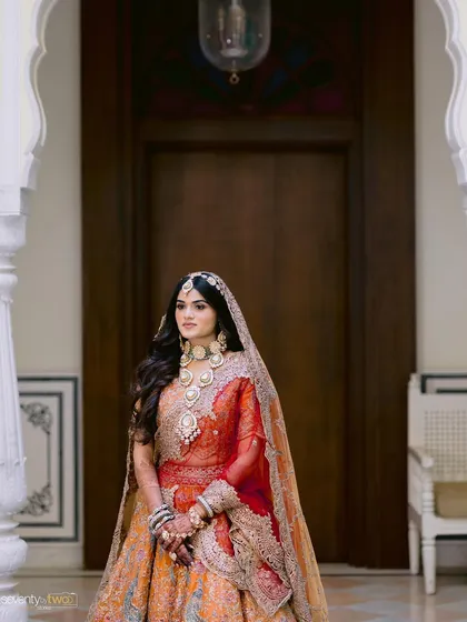 A regal portrait of a bride in her orange and red lehenga, framed by the ornate white arches of a palace in Udaipur.