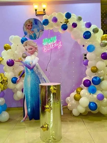 A magical Frozen-themed party for a 4th birthday, featuring an Elsa cutout, a balloon arch in shades of white, blue, and purple, and a neon "Happy Birthday" sign.
