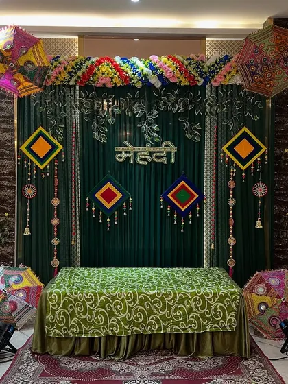 An elegant Mehendi stage with a dark green backdrop, colorful umbrellas, and traditional hangings, perfect for indoor banquet hall events.