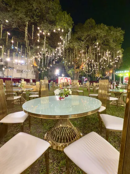 A round guest table with modern gold chairs, set under a canopy of hanging lights for a magical outdoor dining experience.