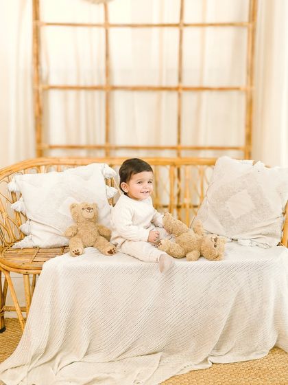 A wider shot of the sitter session, showing the baby comfortably seated on a rattan bench with his teddy bears. The simple, bright setup keeps all the focus on the child.