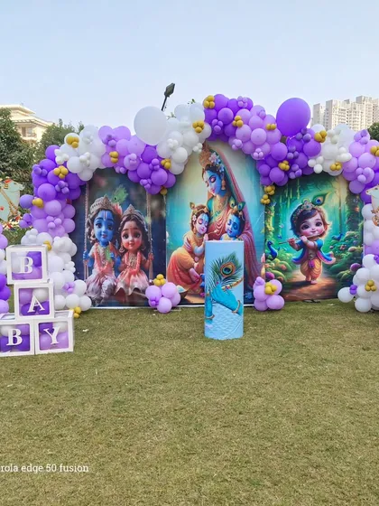 A grand outdoor setup for a baby's function with a Krishna theme, featuring large backdrops and a purple and white balloon arch.