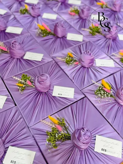 A stunning display of gifts wrapped in pleated lavender fabric. Each package is adorned with a fabric rose knot and a small, colorful bouquet of yellow and pink dried flowers, perfect for spring events or wedding favors.