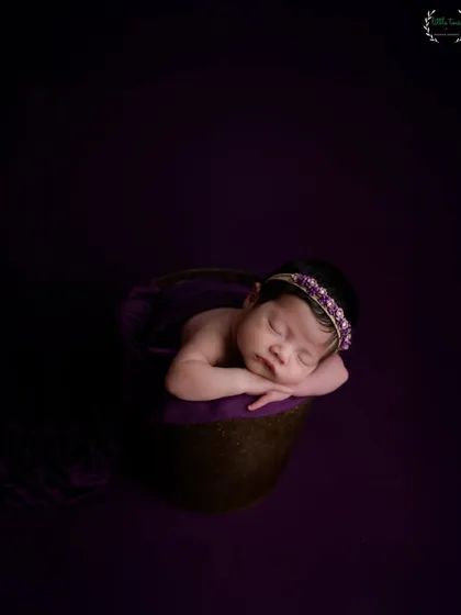 A newborn baby sleeps in a basket against a rich, dark purple backdrop, creating a dramatic and moody portrait. This style offers a beautiful contrast that makes the baby's features stand out.