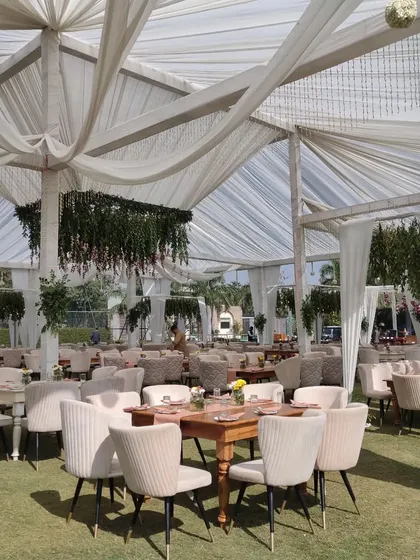 This is a daytime luncheon setup under a large, elegantly draped tent. I arranged the seating with comfortable upholstered chairs and wooden tables to create a sophisticated yet relaxed atmosphere for guests.