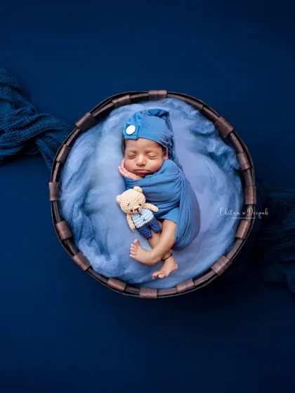 A classic newborn pose, with the baby swaddled snugly in blue and tucked into a rustic basket with a tiny teddy bear. The deep blue background creates a rich, portrait-like effect.