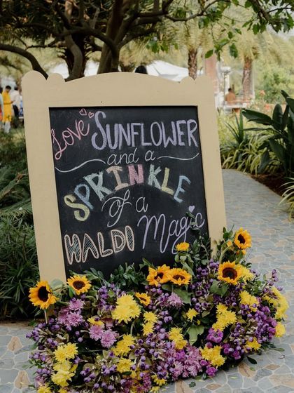 The charming chalkboard welcome sign, with handwritten pastel text and a base of sunflowers and purple blooms.