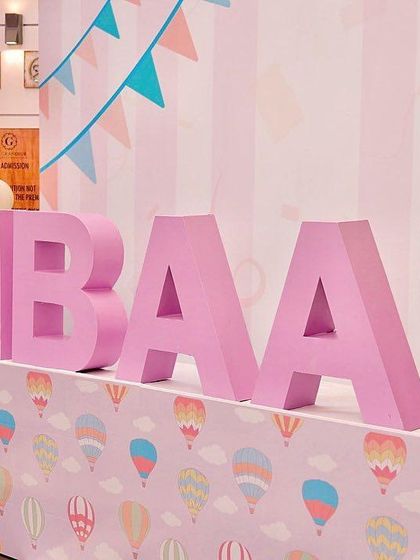 A personalized 3D name sign for "Ibaan" at his pastel carnival party. The large pink letters stand out against a hot air balloon patterned base, next to a whimsical popcorn prop filled with pastel balloons.