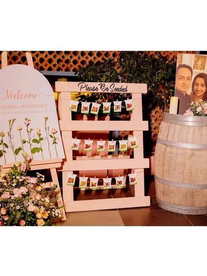 A complete welcome station for an anniversary party. It includes a custom welcome sign, a creative "Please Be Seeded" display with seed packets as favors, and a rustic barrel accent.