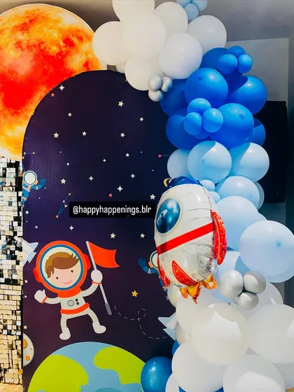 A detailed shot of the space-themed backdrop, featuring a rocket ship foil balloon blasting off alongside a cascade of blue and white balloons, with planet cutouts adding to the cosmic scene.