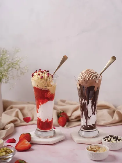 A pair of tall, decadent milkshakes, one strawberry and one chocolate. The styling with fresh fruit and chocolate chips highlights the ingredients and makes the drinks look irresistible.