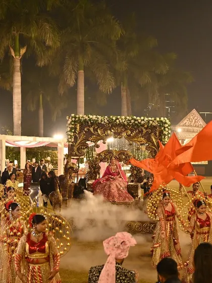A truly grand bride entry. The bride is carried on an ornate palanquin, surrounded by a large troupe of dancers with flags and props, creating a scene of royal splendor.