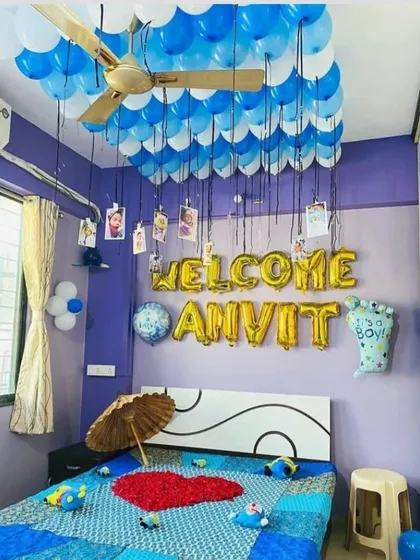 A 'Welcome Anvit' room decoration for a newborn baby boy. The setup includes ceiling balloons with hanging photos, foil letters, and a bed decorated with a rose petal heart.