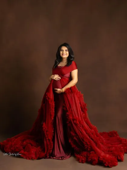 A radiant portrait of an expecting mother in a dramatic red gown. She is wrapped in a ruffled tulle overlay, creating a look of pure glamour against a brown studio backdrop.