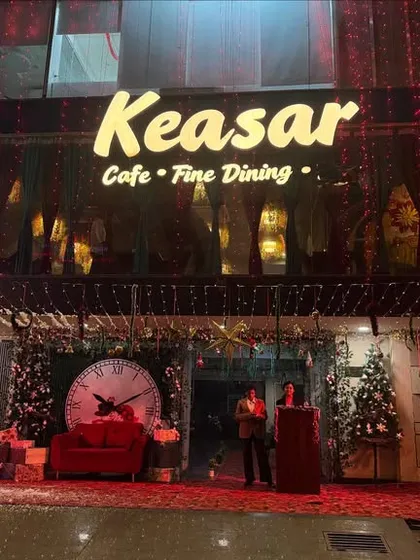 A large-scale Christmas decoration for the entrance of Keasar Cafe. The setup includes a photo booth with a giant clock, a Christmas tree, and gift boxes.