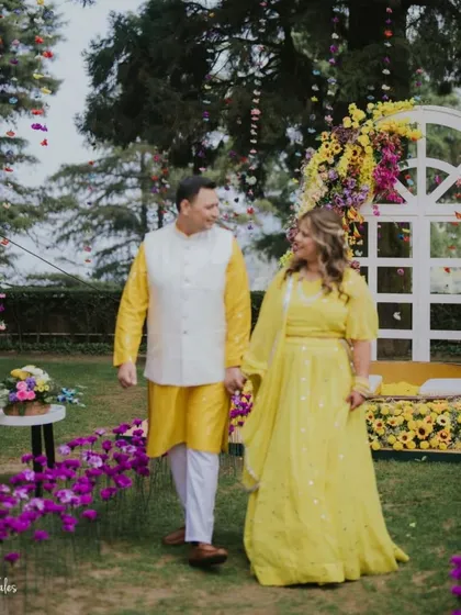 A walk in a garden of sunshine. The couple, dressed in vibrant yellow, strolls through their beautiful Haldi ceremony setup, featuring a floral arch and purple flower beds that create a stunning color contrast.