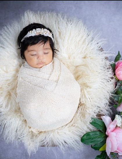 Simplicity and elegance define this newborn portrait. The baby is wrapped in a classic cream swaddle, resting on soft white fur and accented with beautiful pink peonies for a touch of grace.