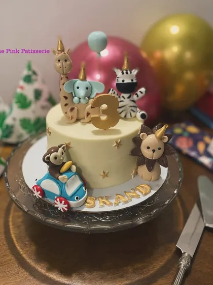 A whimsical party animal cake where a monkey drives a car. This fun design features a giraffe, elephant, zebra, and lion all wearing party hats for a 3rd birthday.