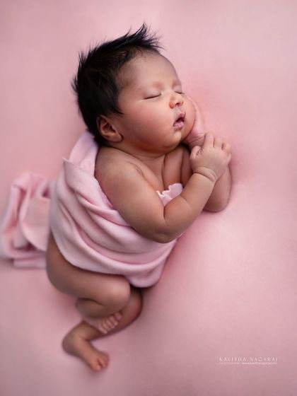 Like a quiet poem in pastels, this one-month-old baby rests her cheeks on her hands. The soft pink background enhances the warmth and innocence of the moment.
