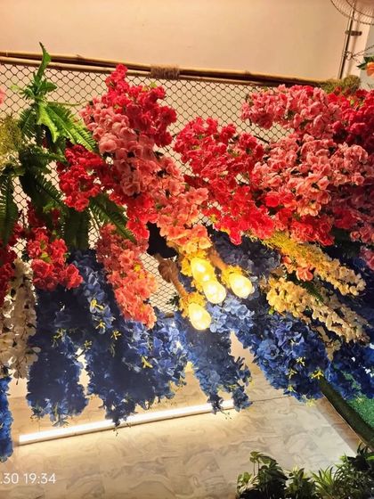 For this project, we created a vibrant ceiling canopy using a wire mesh base. We attached dense clusters of colorful artificial flowers in red, blue, and yellow, weaving in lights for a festive and dramatic effect.