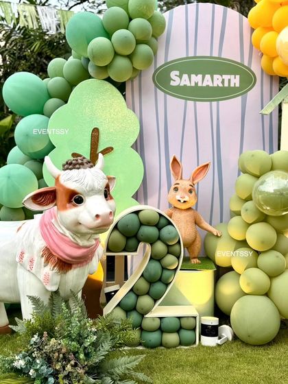 A close-up of the farm animal theme, featuring a friendly cow and rabbit alongside a balloon-filled marquee number "2." The green balloon garland adds a touch of fun to the rustic setting.