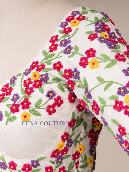 A garden of flowers on a blouse. This white raw silk piece is covered in vibrant, multi-colored floral French knot embroidery, making it a cheerful and eye-catching addition to any festive wardrobe.