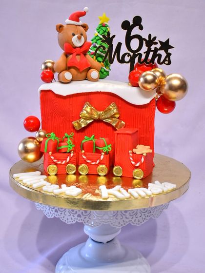 A festive Christmas-themed cake to celebrate a 6-month milestone. The cake is decorated with a teddy bear in a Santa hat, a Christmas tree, and edible presents.