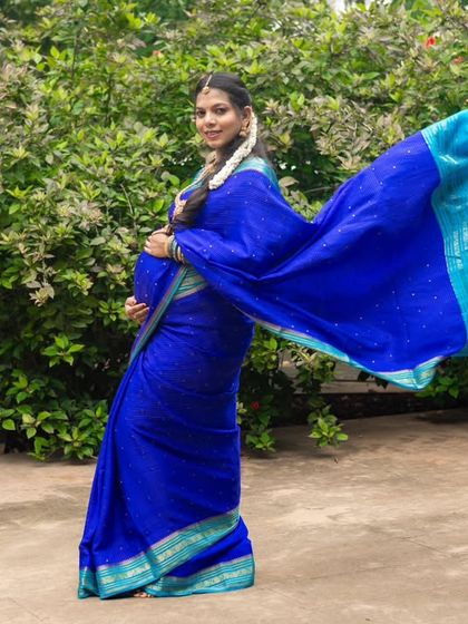 The grace of a traditional saree in motion. This outdoor shot captures the beautiful flow of the royal blue saree against a natural green backdrop, highlighting the elegance of the mom-to-be.