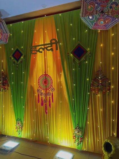 An indoor Haldi setup with green and yellow drapes lit by fairy lights. The decor includes a central dreamcatcher, Rajasthani umbrellas, and decorated matkas for a traditional feel.
