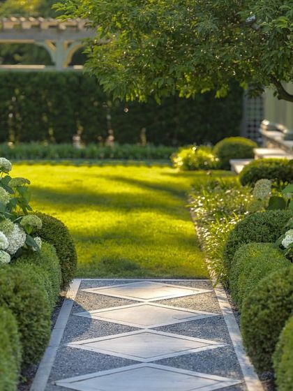 A formal garden path with a diamond pattern created from stone and gravel. The path is bordered by low boxwood hedges and flowering hydrangeas, leading the eye towards a sunlit lawn.