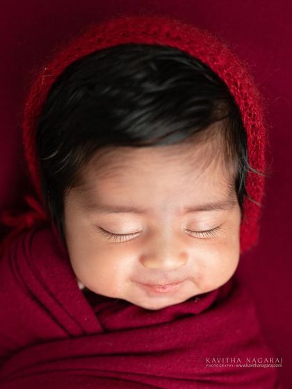 A close-up of a newborn's sweet, smiling face, wrapped in a deep red bonnet and swaddle. This image is all about the pure joy and love a new baby brings.