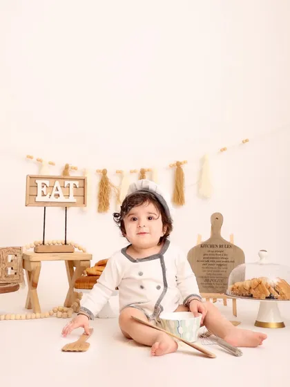 The baby chef sits amidst his baking setup, looking directly at the camera. This full-view shot showcases the entire charming and rustic-themed environment.