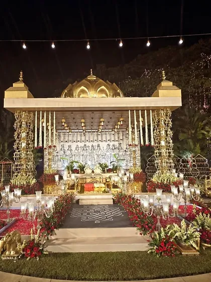 An opulent wedding mandap designed for an outdoor amphitheater. The structure features our custom-made brass pillars, a domed roof, and is surrounded by lush red florals and traditional lamps, all under a canopy of twinkling firefly lights.