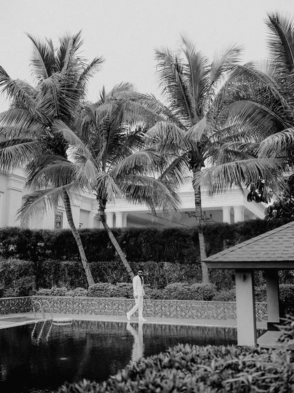 A minimalist black and white shot of the groom by the pool. The reflection in the water and the strong lines of the palm trees create a stylish and artistic composition.