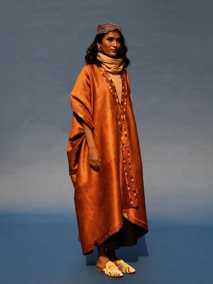 A full-length shot of a flowing, rust-colored kaftan. The simplicity of the pose and the solid background allow the garment's elegant drape to take center stage.