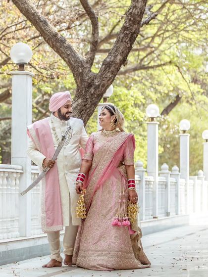 A classic portrait of a Sikh couple, with the groom holding his kirpan. Their coordinated pink and white outfits look stunning against the clean, architectural background.