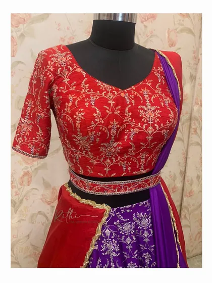 A close-up of the half-saree's blouse and waistband. The red blouse features intricate all-over embroidery that contrasts beautifully with the purple brocade skirt.