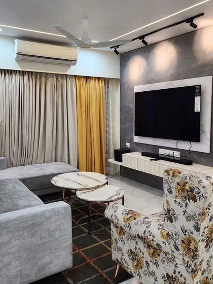 This cozy corner of a living room shows a comfortable grey L-shaped sofa paired with a floral accent chair. The feature wall behind the TV combines a dark textured panel with light wood fluting, illuminated by track lights.