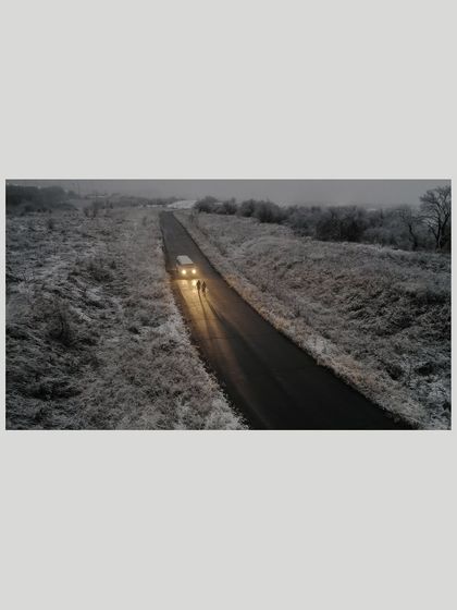 An aerial view of a couple walking down a desolate road in Almaty, illuminated by car headlights. This shot creates a dramatic, cinematic mood for a pre-wedding film.