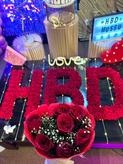 A close-up of the romantic details of a birthday celebration, including a bouquet of red roses and "HBD" spelled out in flowers on the table.