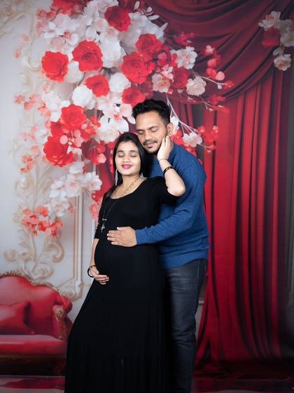 An expecting couple shares a loving embrace in front of a bold red floral backdrop, with the mother-to-be in a classic black dress.