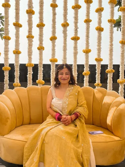 The bride is all smiles, seated on a beautiful yellow sofa against a backdrop of floral strings.