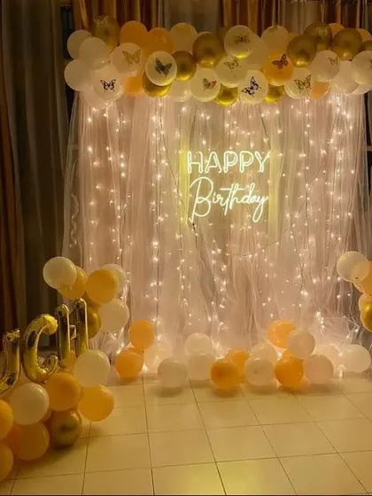 A simple yet elegant birthday setup with a fairy light curtain backdrop and a gold and white balloon garland.