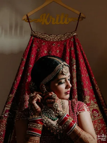 A beautiful shot of the bride adjusting her earring, with her red lehenga and personalized 'Kriti' hanger in the background.