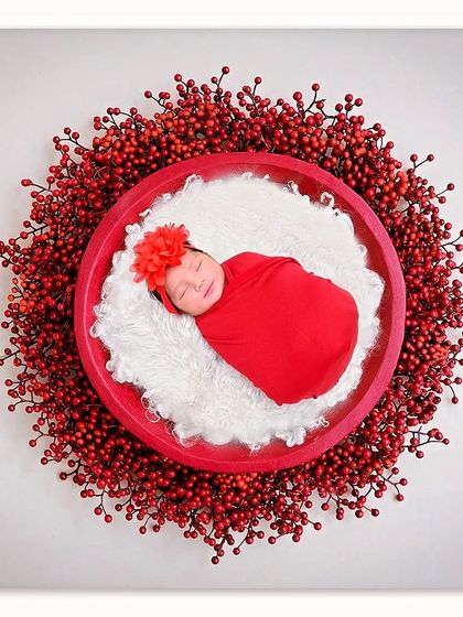 A bold and beautiful top-down shot. The baby is wrapped in bright red and placed in the center of a wreath made of red berries.
