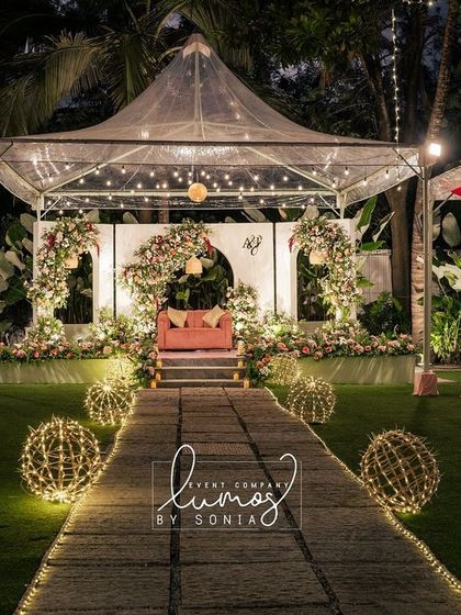 An aesthetic outdoor wedding dinner setup under a clear tent. The path is lined with glowing orbs, leading to a beautifully decorated stage with pink and white florals.