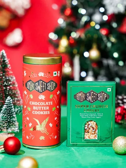 A product shot of Bombay Sweet Shop's Christmas collection, featuring their signature cookies and chikki. The simple, festive styling with mini trees makes the colorful packaging pop.